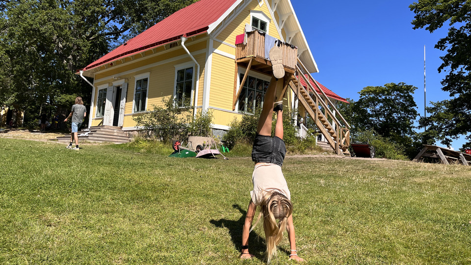 Ett barn står på händer på en gräsmatta framför ett gult trähus.