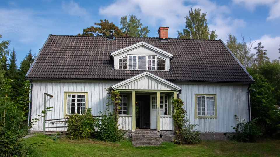 Ett vitmålat trähus i två plan. Veranda vid entrén. Trädgård med gräs, buskar och träd framför huset.
