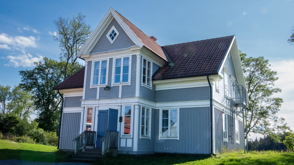 Ett blått trähus i två plan med stora fönster. Runtom syns gräsmattor, buskar och träd.