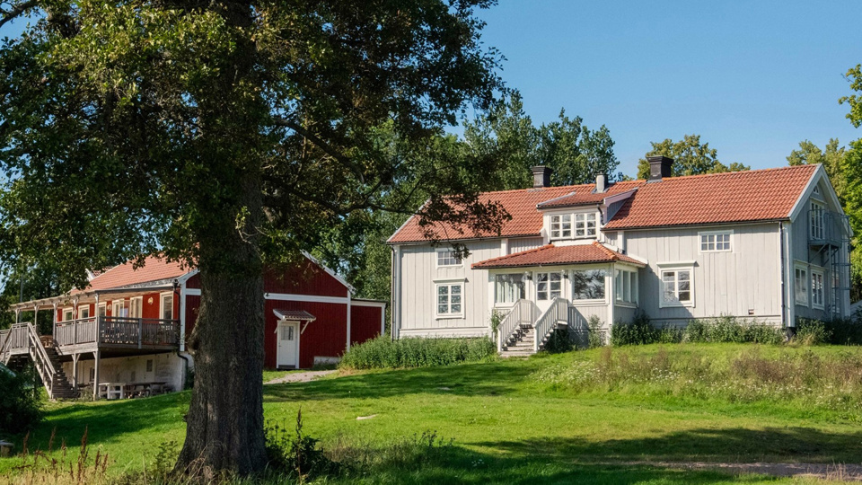 Ett ljust trähus i två plan. En trappa leder upp till entrén. Trädgården är lummig med gräs, blommor och buskar. Till vänster ett rött trähus.