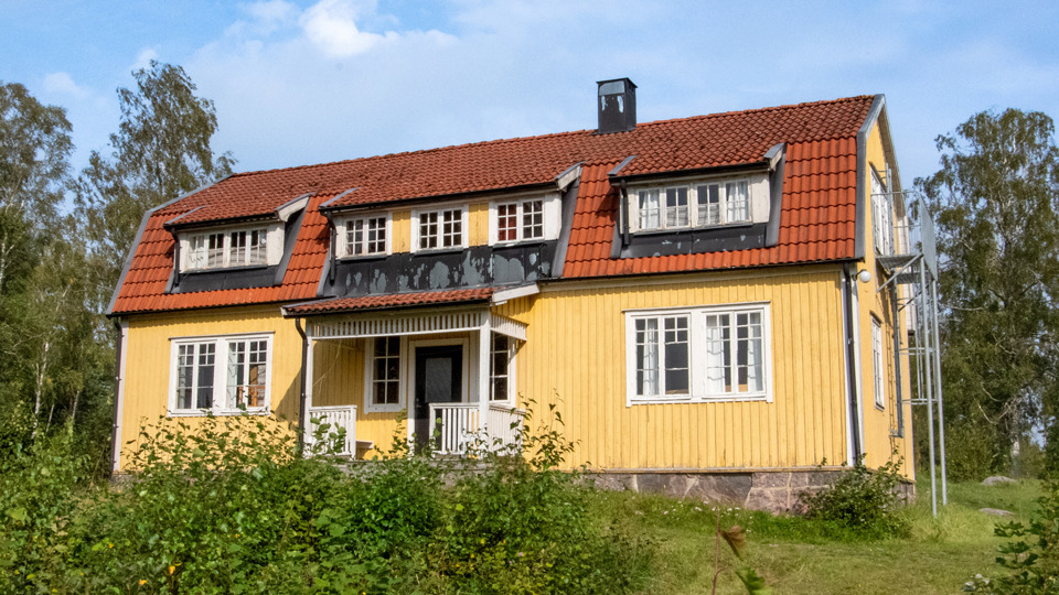 Ett stort gulmålat trähus i två plan. Veranda vid entrén och en gräsplan och buskar framför huset.