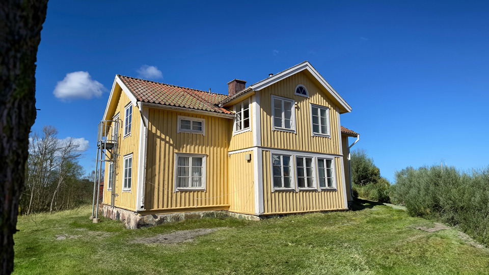 Ett gult trähus i två plan, högt beläget och omgivet av gräsmatta och träd i en sluttande backe.