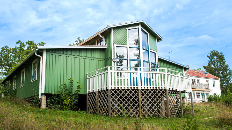 Ett grönt trähus med veranda. Bakom syns ett vitt hus. Gräsmattor och träd.