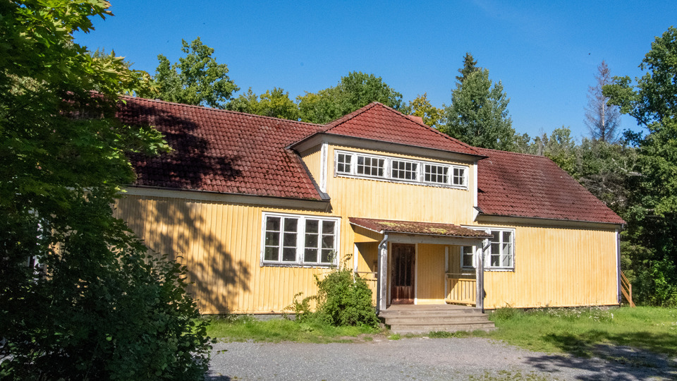 Ett stort gult trähus med liten veranda. Framför huset en gräsmatta och grusplan. Bakom huset buskar och träd.