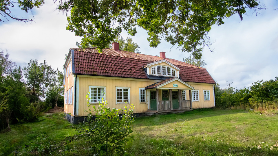 Ett gult trähus i två plan med veranda vid entrén. En lummig trädgård med gräs, buskar och träd.