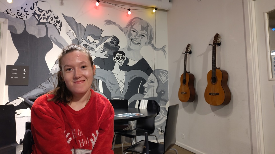 En glad tjej sitter i ett rum med stolar, bord och gitarrer upphängda på väggarna.