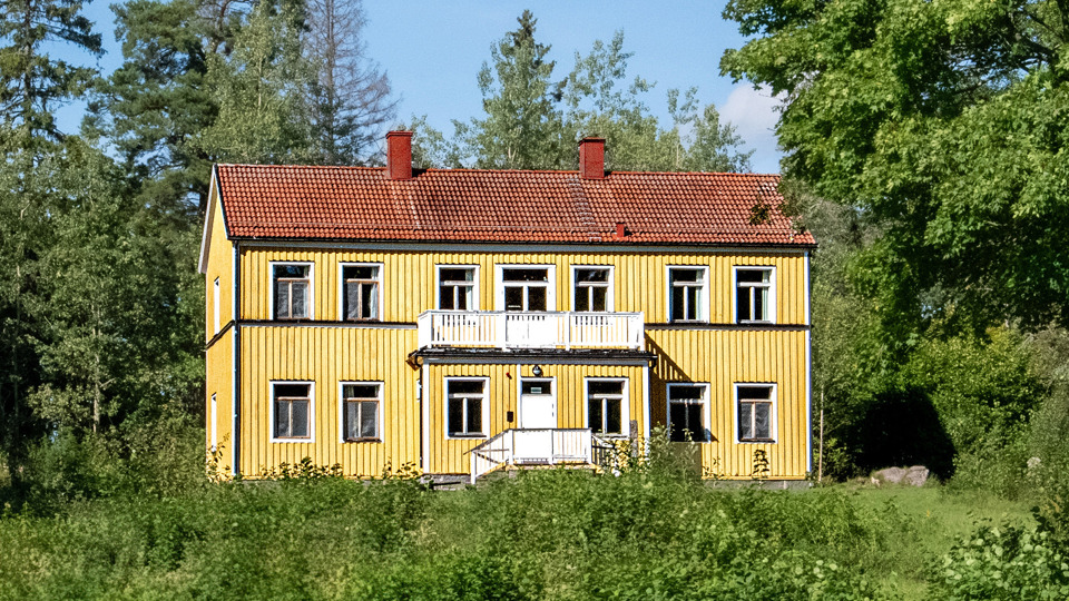 Ett stort gult trähus i två plan med verandor. Buskar, träd och gräsmatta runt huset.