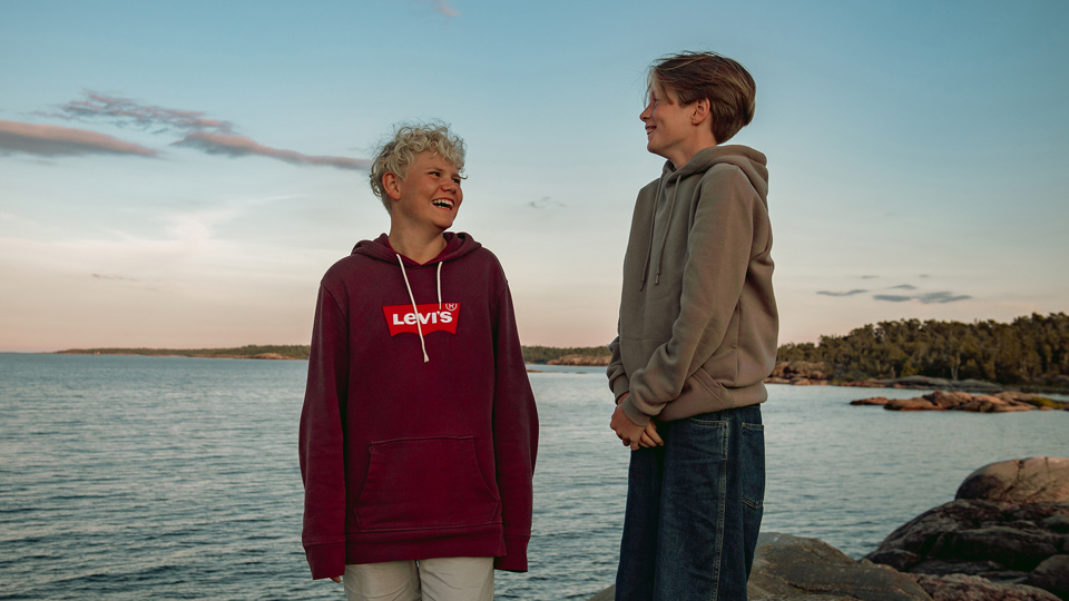 Två barn står på klippor nere vid vatten i svagt solljus. De skrattar tillsammans.
