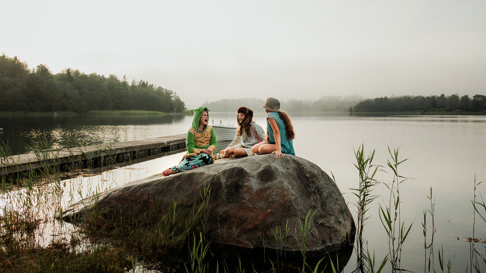 Tre barn sitter på en klippa vid vattnet. I bakgrunden är det lite dimmigt över skogen.