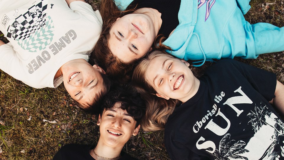Fyra leende ungdomar ligger med sina huvuden mot varandra i gräset. De kisar mot solen.