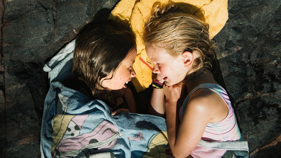 Två barn som nyss har badat ligger på en klippa i solen, skrattar och tisslar tillsammans.
