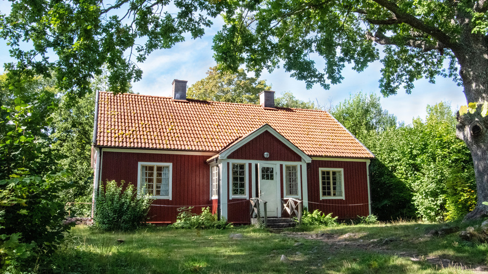 Ett rött mindre trähus omgivet av gräsmatta och lummiga träd