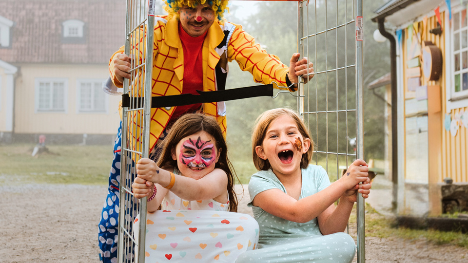 Två barn sitter i en rullvagn som körs av en man som är utklädd till clown.