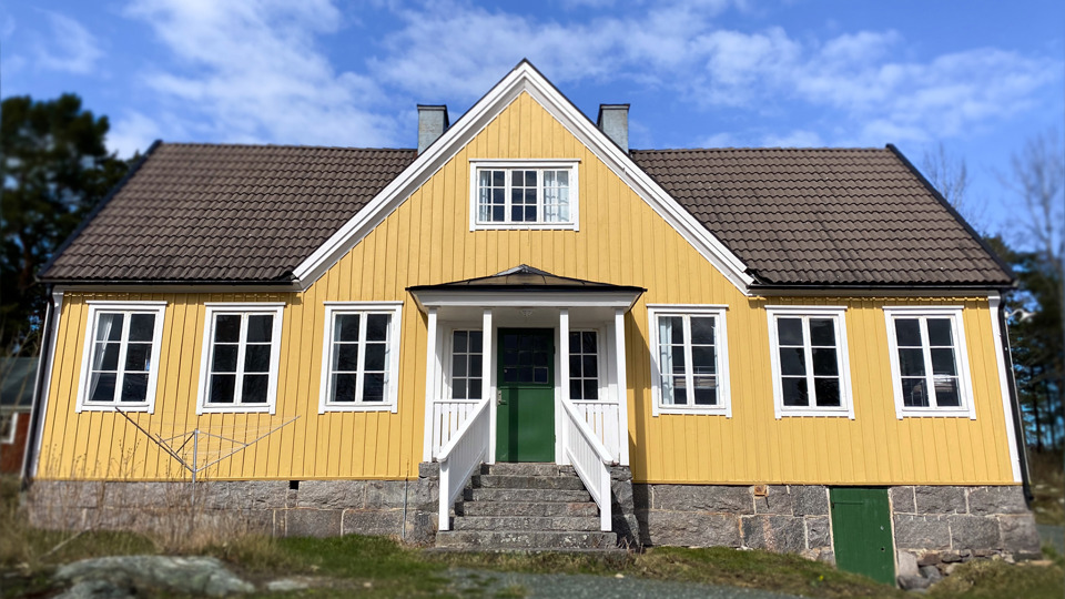 Ett gult trähus i två plan. Framför huset skymtar en gräsmatta, bakom huset skymtar träd.