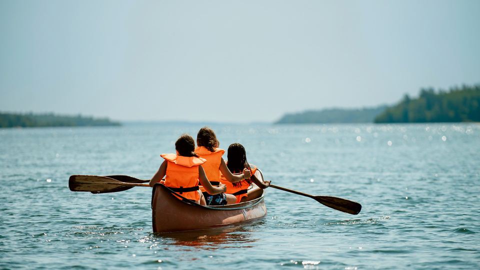 Tre barn sitter i en stor kanot på sjön och paddlar.