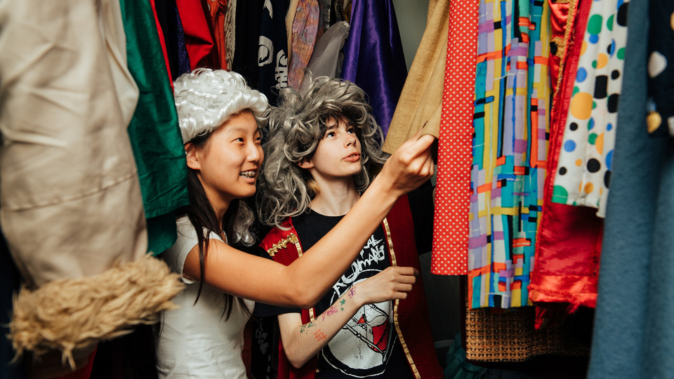 Två barn iförda peruker står bland en massa utklädningskläder i en stor garderob.