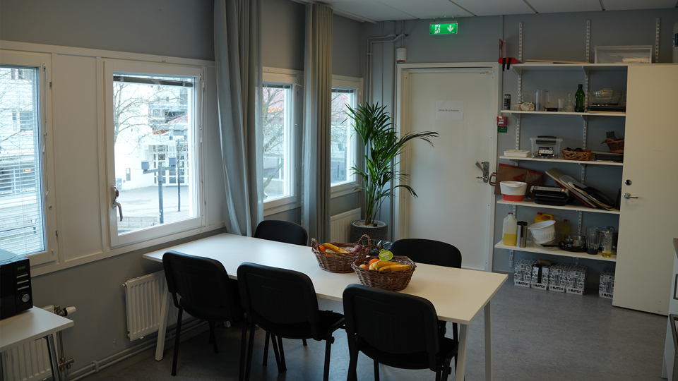 Ett kök. På ena väggen sitter flera hyllor som det står redskap och material på. Vid fönstret står ett matbord med sex stolar omkring, foto