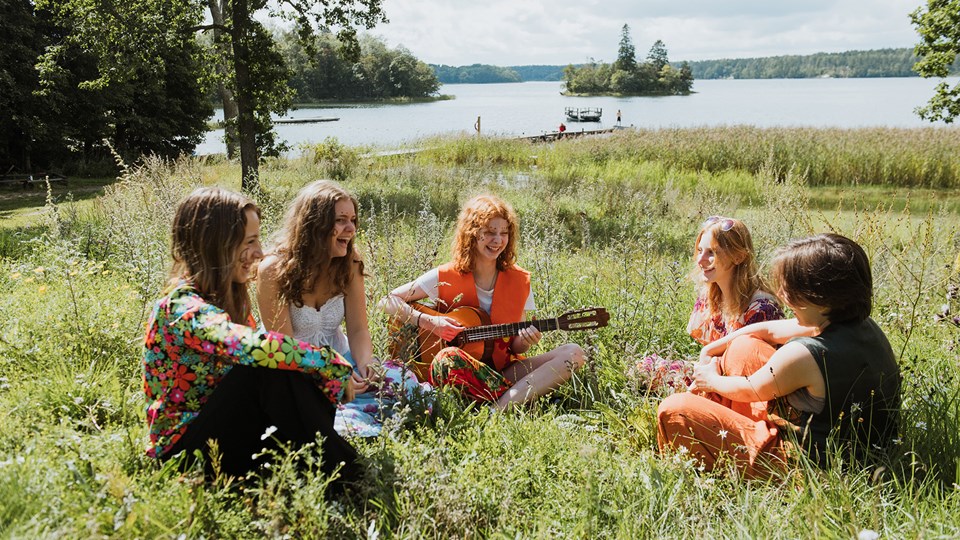 Fem barn sitter i gräset och sjunger och spelar gitarr.