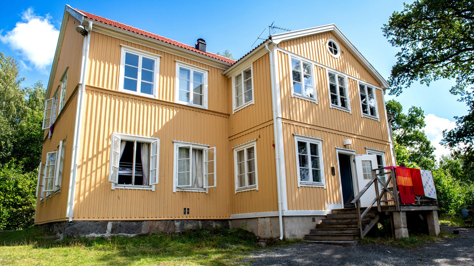 Ett gult trähus i två plan. Veranda vid ingången. Trädgård med gräsmatta, träd och en grusgång.