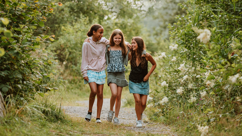 Tre flickor går i bredd och håller om varandra på en stig i skogen.