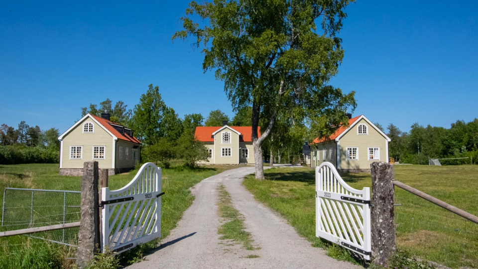 En vit trädgrind i förgrunden som leder in till tre ljusa trähus i två plan runt en gårdsplan. Gräsmattor och träd.