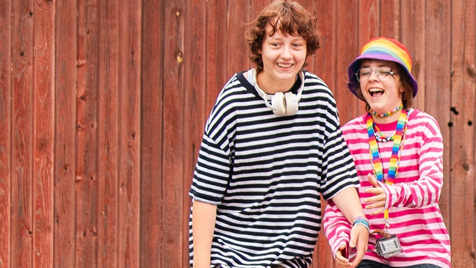 Två barn står framför ett rödmålat plank. De skrattar och håller varandra i handen.