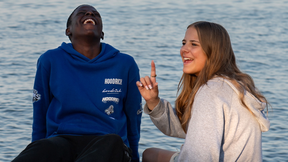 En pojke och en flicka sitter på klippor vid vatten. De skrattar och skojar med varandra.