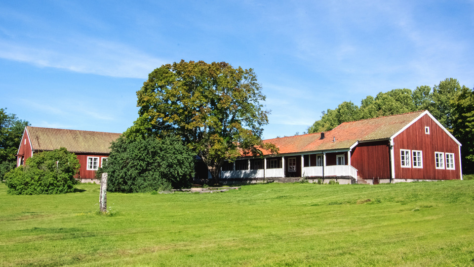 Ett stort rött trähus i vinkel. En lång veranda längs en sidan och en stor gräsmatta framför huset.