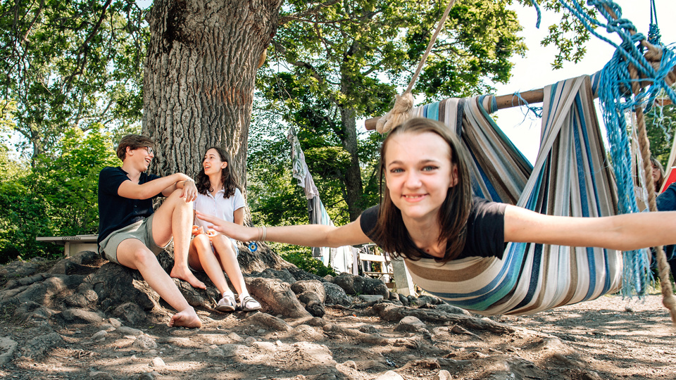 Två barn sitter vid ett träd och ett barn ligger i en hängmatta och gungar.