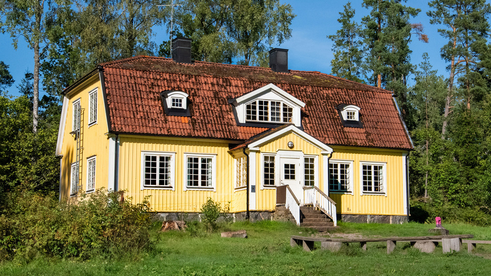 Ett stort gult trähus i två plan. En trädgård med sittbänkar och gräsytor. Bakom huset ser man träd.