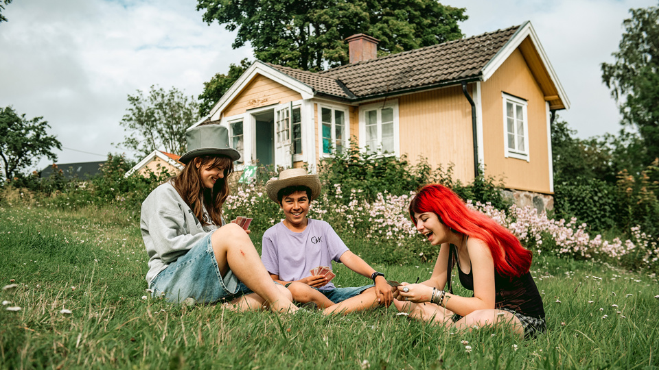 Tre ungdomar sitter i gräset och spelar kort. Bakom dem ett gult trähus och blommande buskar.