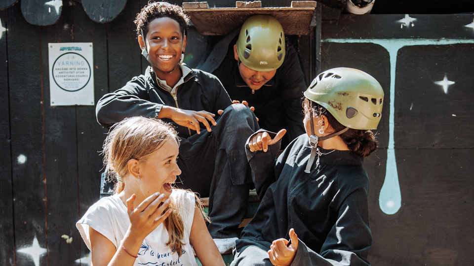 Fyra barn sitter i en trappa utomhus och skrattar tillsammans. Två av barnen har klätterhjälmar på sig.