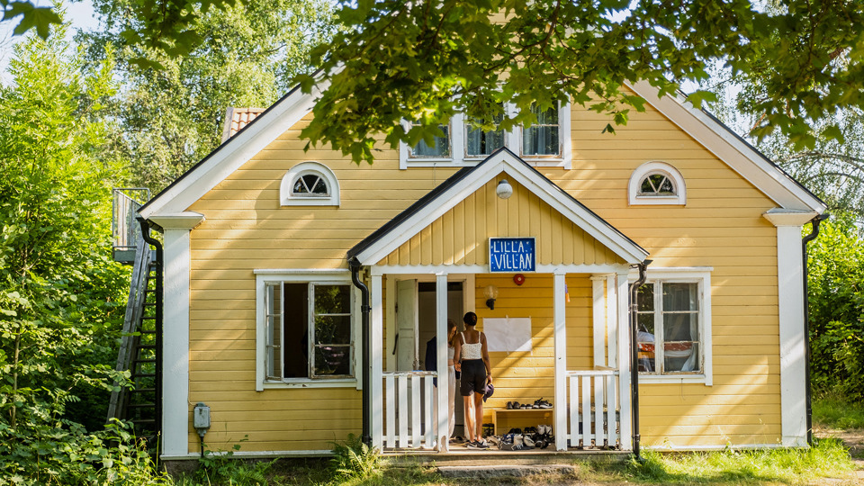 Ett gult trähus med veranda och skylt "Lilla villan". Inbäddat i grönska.