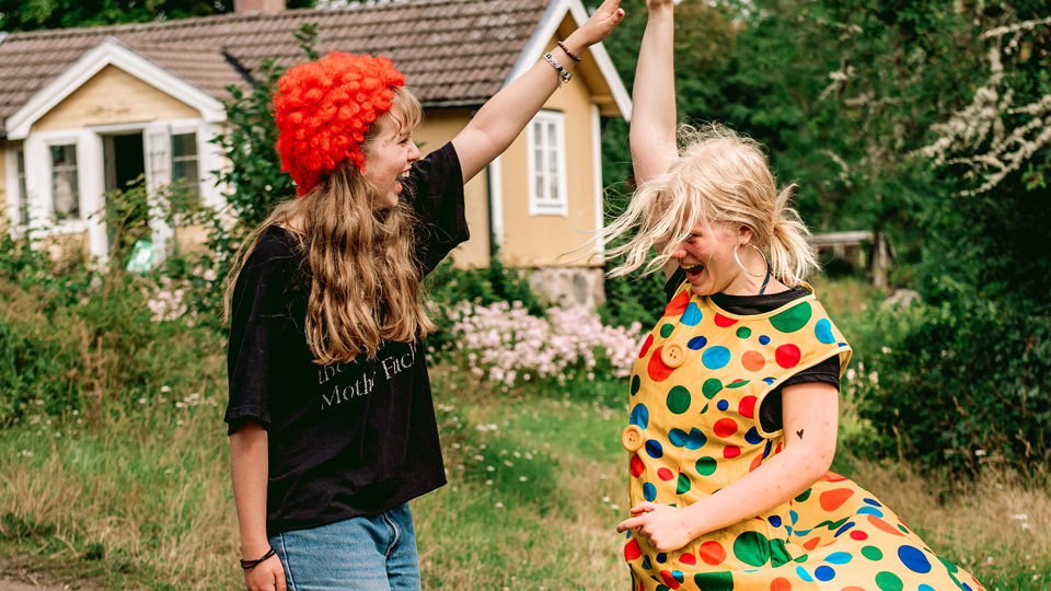 Två barn står utomhus framför ett gult hus. De skrattar och klappar händerna mot varandra. De är utklädda i röd peruk och prickig klänning.