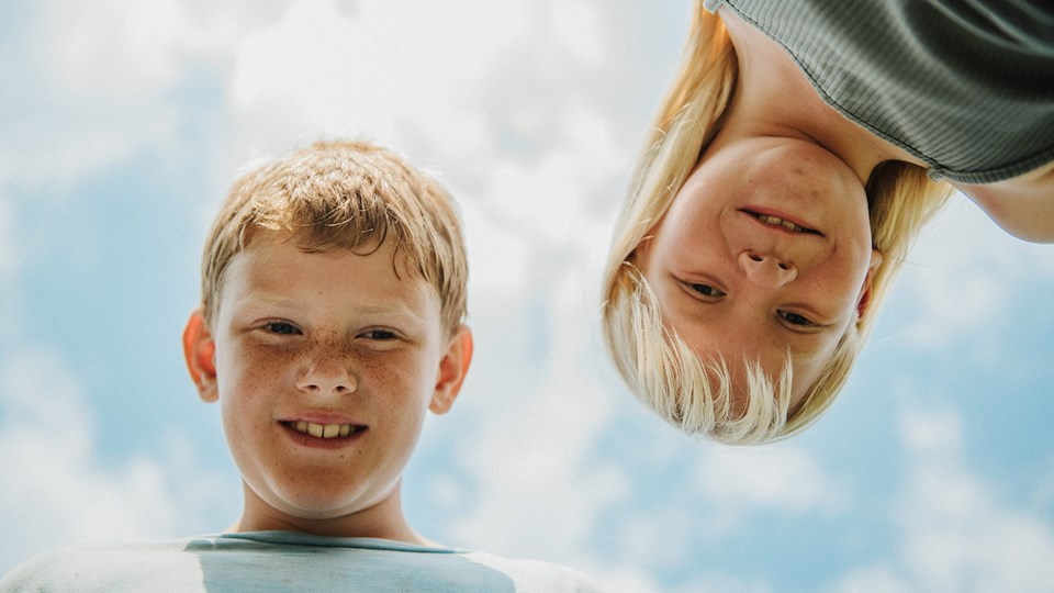 Två barn ler och tittar ner mot kameran med himlen som bakgrund.