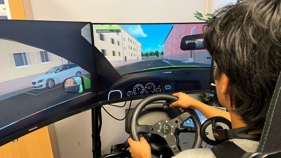 En tonårig pojke sitter i en körsimulator med bilbälte på sig. Framför sig har han två dataskärmar och en ratt, foto