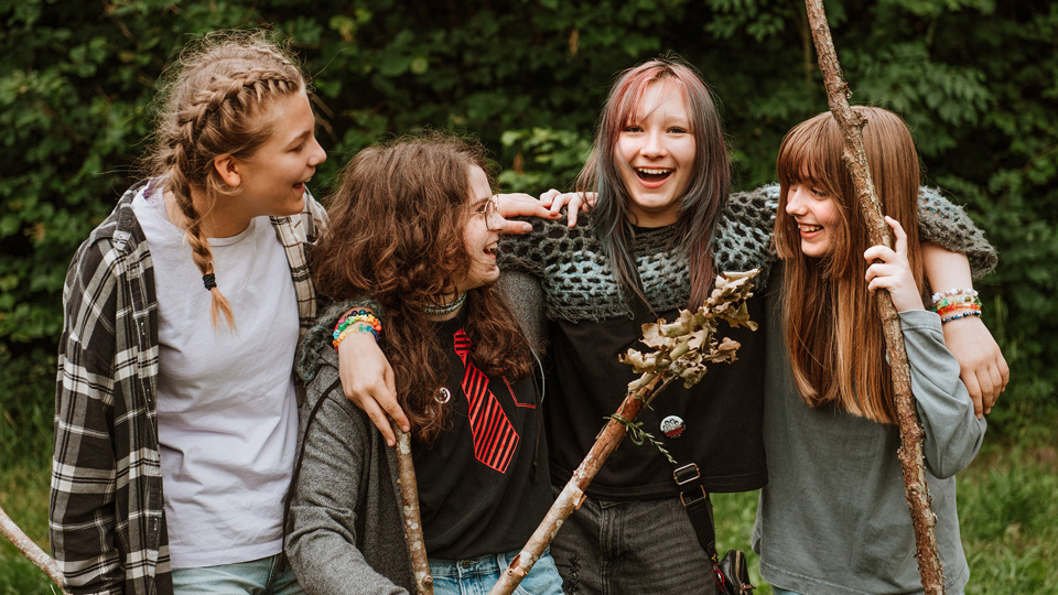 Fyra glada barn står på rad utomhus och håller om varandra. I handen håller de långa pinnar.