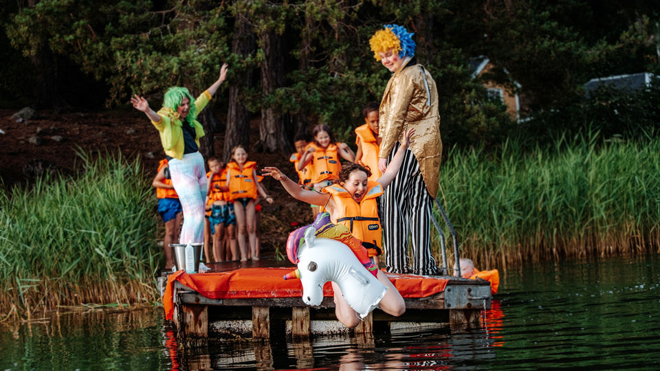 En grupp med barn leker på en brygga. Ett barn hoppar i vattnet och två utklädda kolloledare hejar på.
