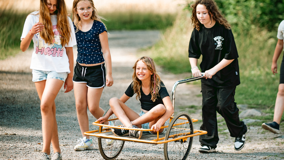 Ett barn sitter på en cykelskrinda och dras fram av ett annat barn. Två barn går bredvid.