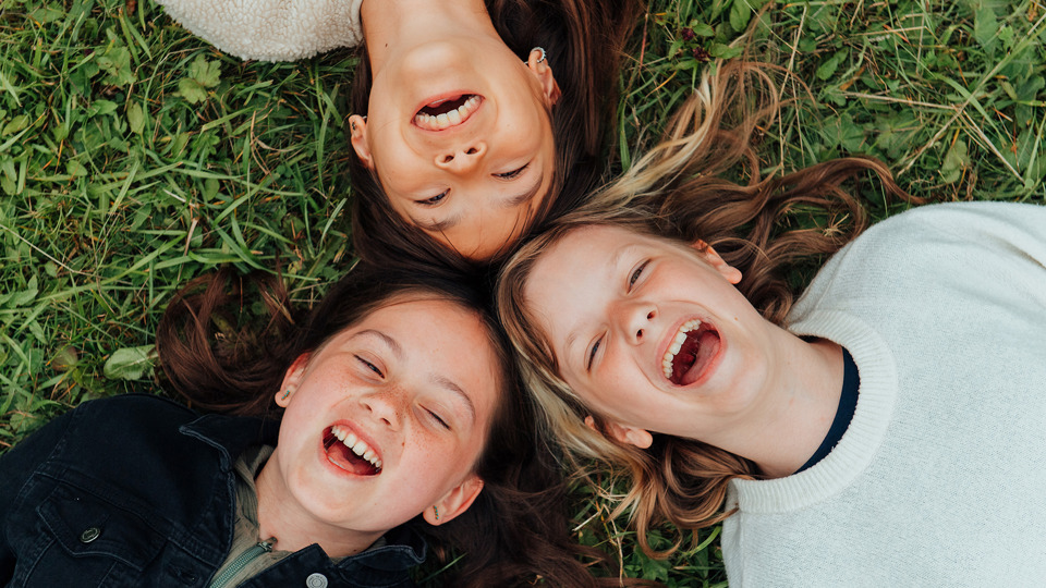 Tre barn ligger i gräset med huvudena mot varandra. De skrattar.
