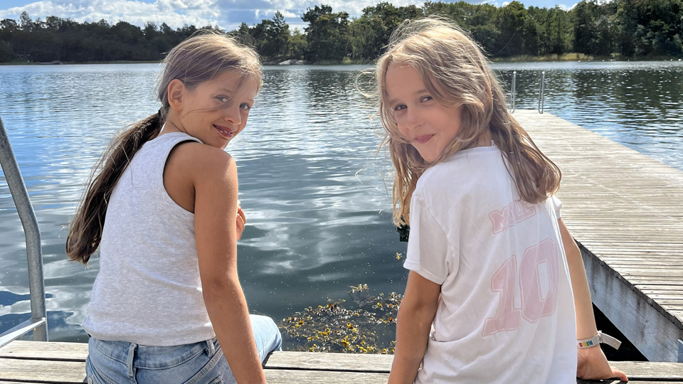 Två barn sitter på en brygga, vänder sig om och ler in i kameran.