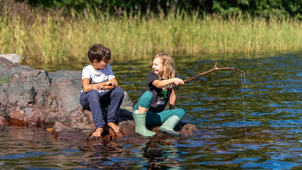 Två barn sitter på en klippa vid en sjö och metar.