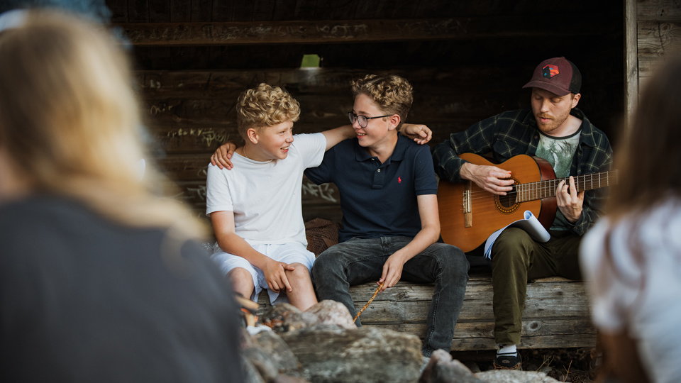 Barn sitter vid en lägereld och grillar. En kolloledare spelar gitarr.