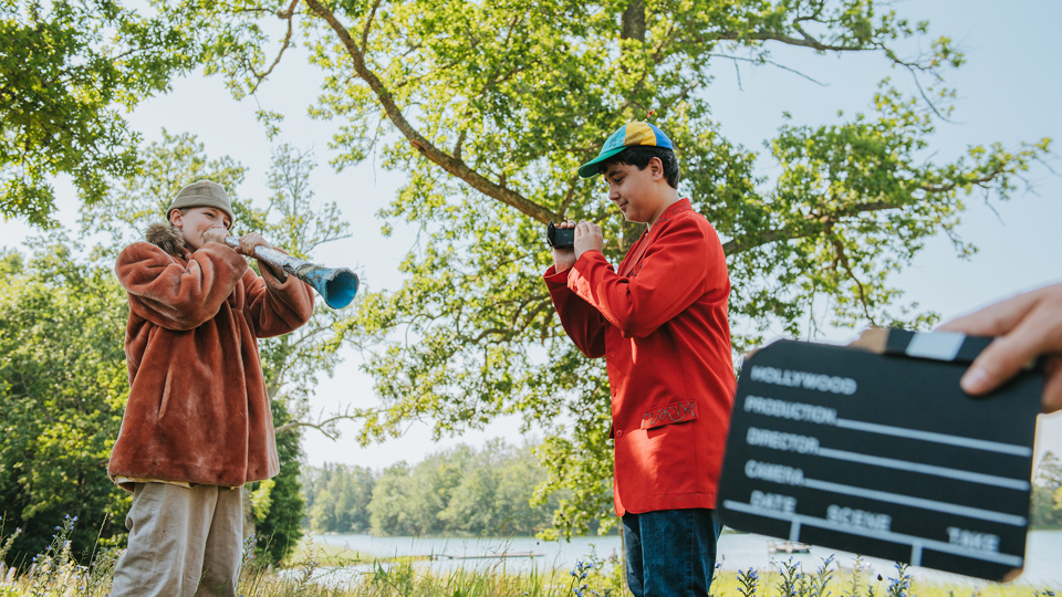 Två barn blåser i musikinstrument och spelar in en filmscen utomhus. I förgrunden syns en filmklappa.