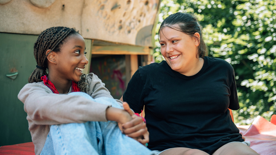 Två barn sitter tillsammans utomhus och ler mot varandra.