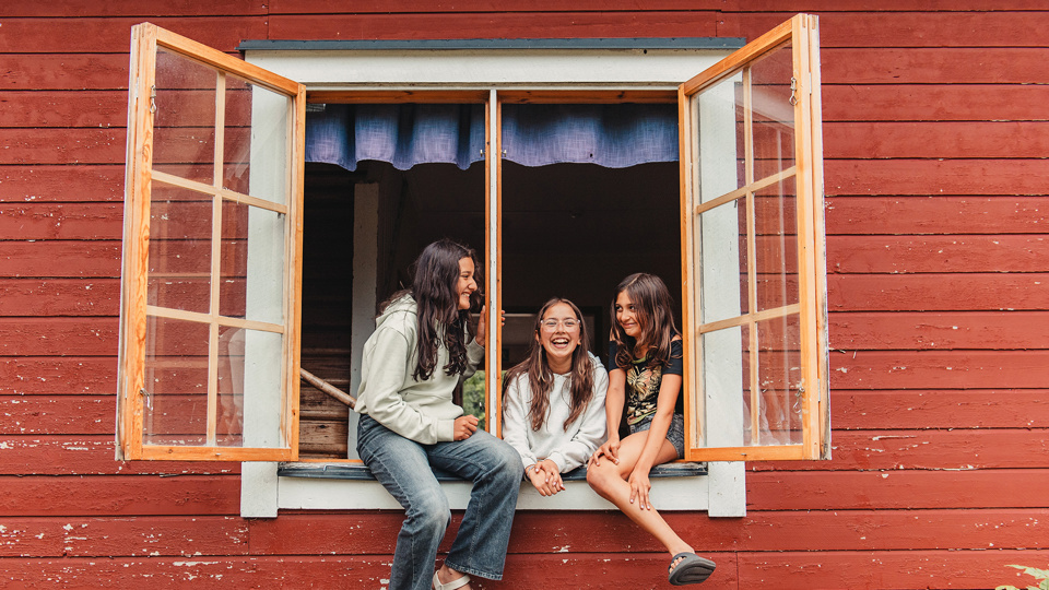 Tre barn sitter på karmen vid ett öppet fönster på ett rött trähus.