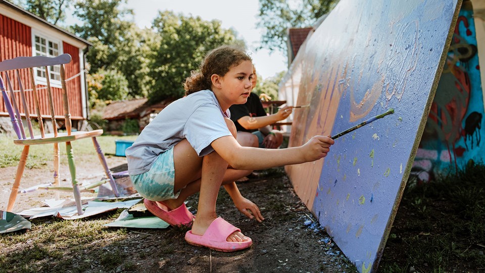 Barn sitter utomhus vid ett plank och målar med pensel.
