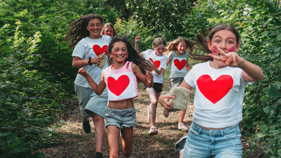 En grupp barn springer i skogen. De har på sig tröjor med stora röda hjärtan.