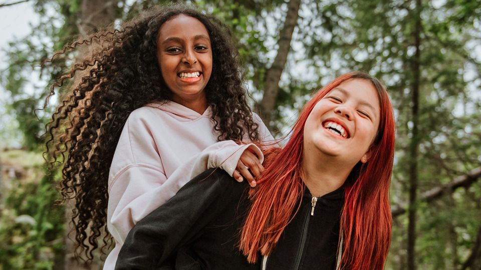 Två skrattande flickor utomhus. Den ena flickan bär den andra flickan på ryggen.