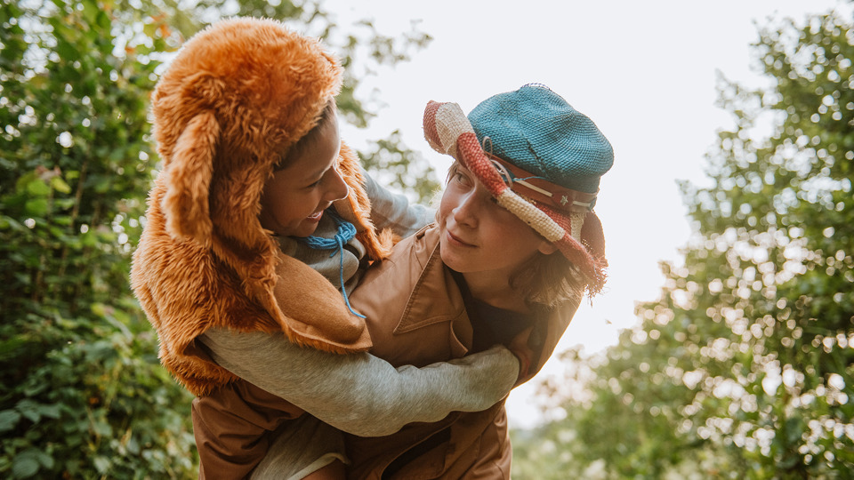 Två barn som är utklädda leker utomhus. Det ena barnet bär det andra på sin rygg.